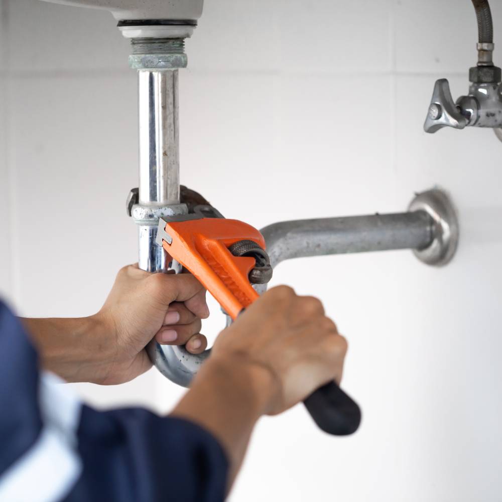 A plumber fixes a pipe fitting under a sink using a pipe wrench.