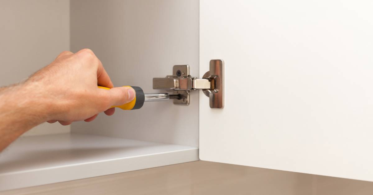 A man uses a yellow screwdriver to attach a plated side-mount hinge inside an empty white kitchen cabinet.
