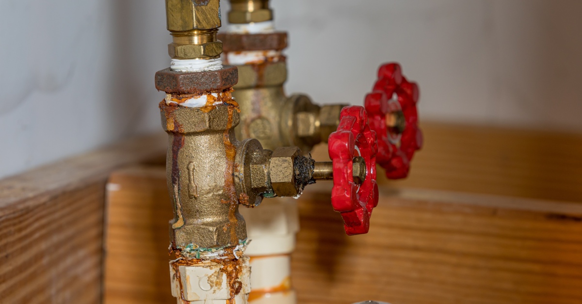A corroded home water value with copper pipes, brass fittings, and a red knob with wooden planks in the background.