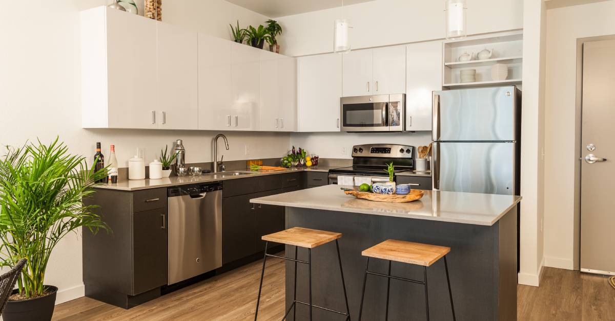A modern apartment kitchen with dark grey bottom cabinets, white top cabinets, stainless steel appliances, and an island.