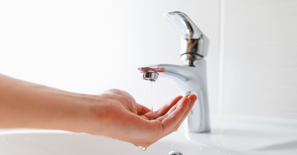 A person holding their hand under a running sink faucet with lower water pressure in a white bathroom.