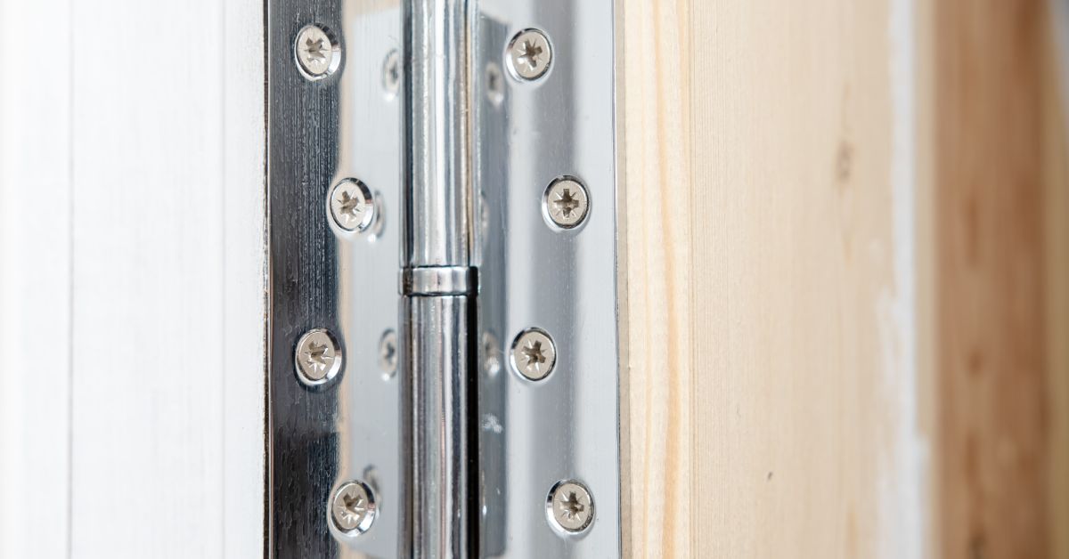 A close-up of a shiny metal door hinge with four screws on each leaf, joining a white door to an unstained surface.