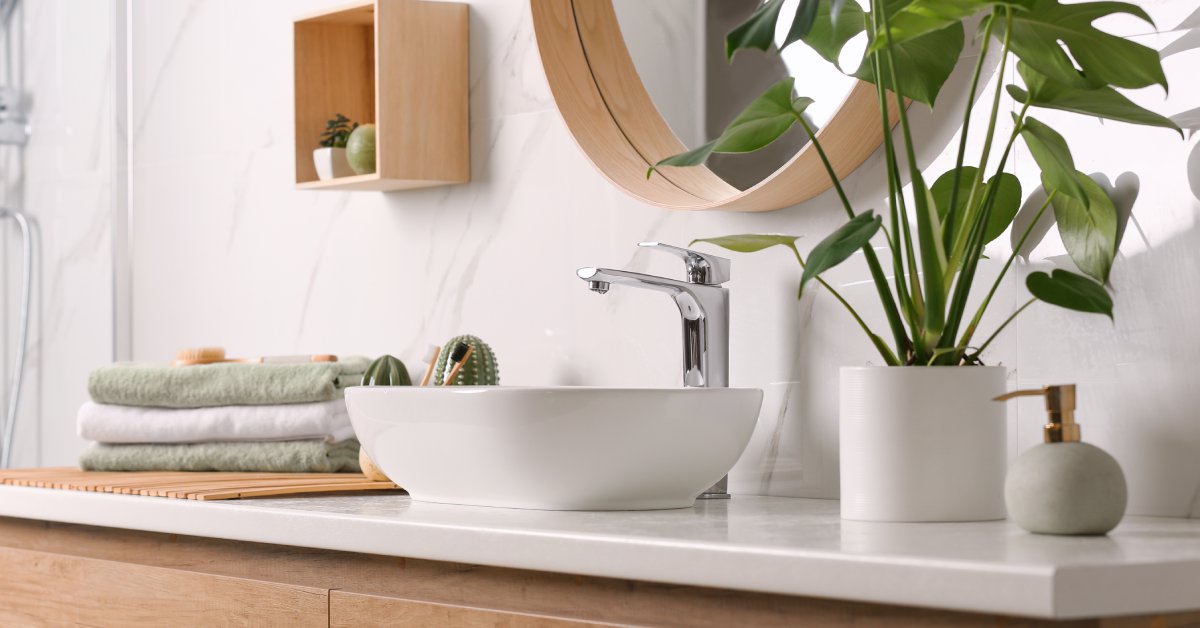 A modern bathroom counter with a vessel sink, stack of towels, and potted plant. A circular, framed mirror hangs on the wall.