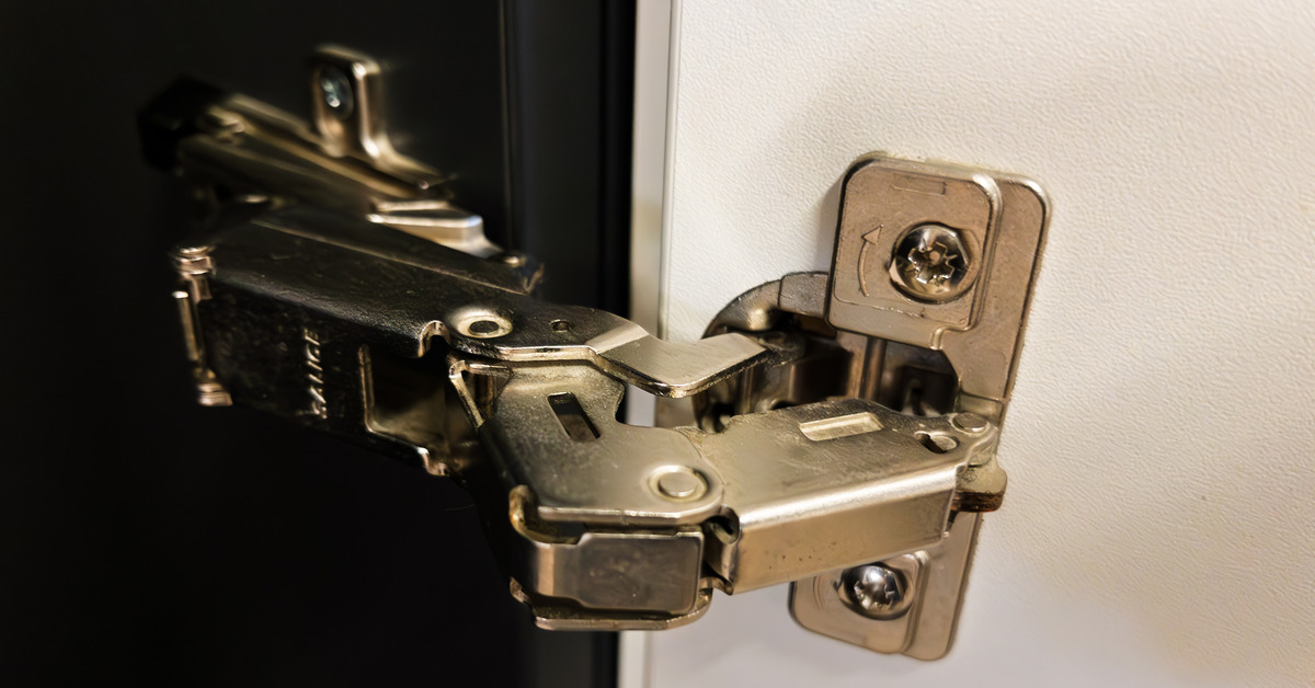 A close-up of a metal soft-close hinge on the inside of a kitchen cabinet. The main cabinet is black, and the door is white.