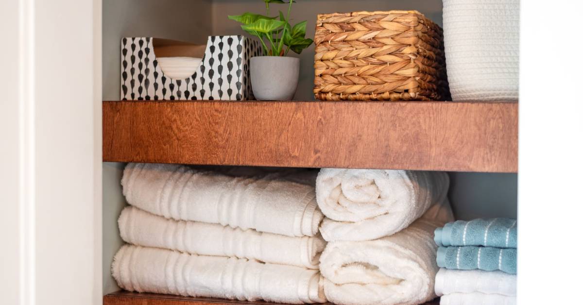 The inside of a bathroom cabinet with thick wood shelves stacked with rolled towels, tissues, and baskets.