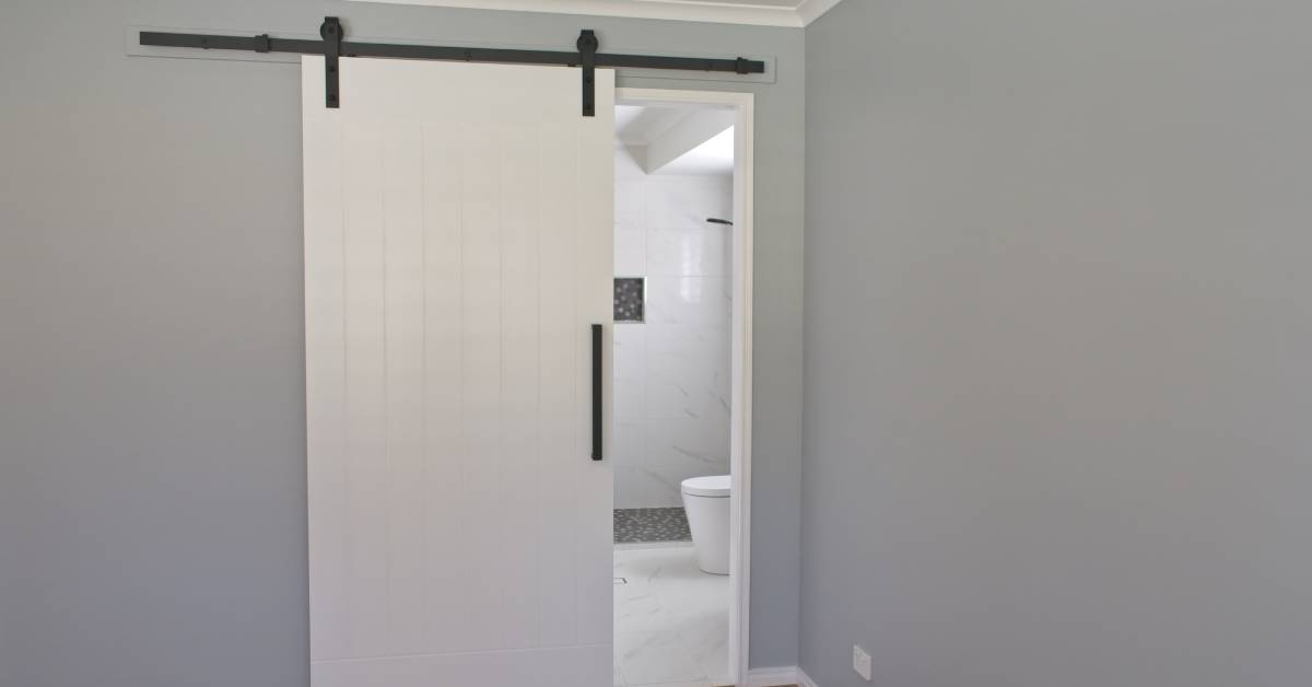 A white sliding barn door on a black track opens from an empty bedroom with grey walls to a bathroom with white tile.